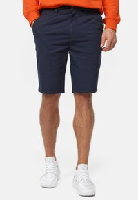 Hose in Marineblau, tailliert, aus einem Baumwollmischgewebe, mit seitlichen Taschen und einem Knopfverschluss, kombiniert mit weißen Sportschuhen.