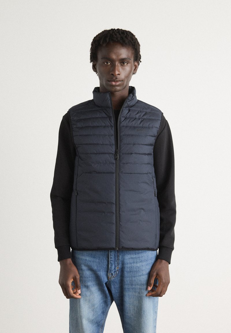 Colete acolchoado azul-marinho com gola alta e fecho de correr. Apresenta costura horizontal, bolsos laterais e um design leve.