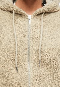 Κοντινή λήψη από ένα μπεζ φλις hoodie με φερμουάρ και κορδόνια σε ασημί χρώμα, που φοριέται πάνω από μια λευκή μπλούζα.