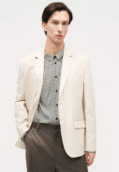 Άνδρας με μπεζ σακάκι, ασπρόμαυρο καρό πουκάμισο και σκουρό καφέ παντελόνι, στέκεται μπροστά σε απλό ανοιχτό φόντο.