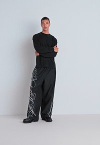 Camisa negra de manga larga combinada con pantalones holgados negros que presentan patrones abstractos en blanco. El modelo está de pie con los brazos cruzados, usando zapatos negros.