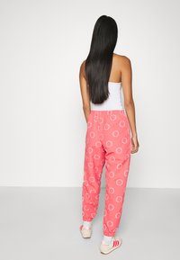 Donna con lunghi capelli neri che volta le spalle, indossando una canotta bianca, pantaloni da jogging corallo con motivi circolari bianchi, calze bianche e scarpe da ginnastica beige.