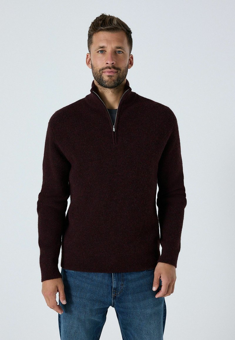 Burgunderfarbener Pullover mit Reißverschlusskragen aus weich gestricktem Stoff, mit langen Ärmeln und lockerer Passform, kombiniert mit blauen Denim-Jeans.