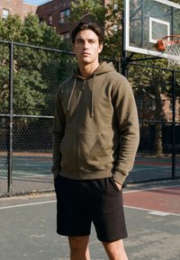 Jeune homme portant un sweat à capuche vert zippé et un short noir, debout sur un terrain de basket en plein air, les mains dans les poches.