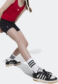 Obuwie charakteryzuje się czarno-kremowym wzorem z trzema czarnymi paskami, zestawione z białymi skarpetami z czarnymi paskami oraz czarnymi krótkimi spodniami sportowymi.