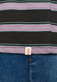 Zwarte t-shirt met korte mouwen, voorzien van horizontale roze en blauwe strepen, met een rode label met "REV" aan de onderrand. Gecombineerd met blauwe denim jeans.