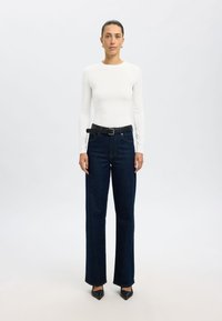 Valkoinen pitkähihainen paita, joka on yhdistetty tummansinisiin korkeavyötäröisiin farkkuihin, joissa on musta vyö. Farkuissa on suora leikkaus ja hienovaraiset ompeluyksityiskohdat.