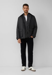 Giacca nera impermeabile con chiusura a zip, due tasche frontali, indossata sopra un maglione a fantasia, abbinata a pantaloni scuri e sneakers bianche.