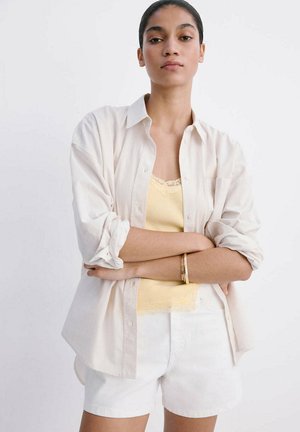 Ung kvinde står med armene over kors, iført en cremefarvet skjorte med knapper over en gul blonder-trimmet camisole og hvide shorts mod en ensfarvet baggrund.