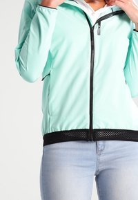 Chaqueta de color verde azulado claro con cremallera frontal, ribete de malla negra en el dobladillo y mangas largas. Presenta una textura suave y un diseño deportivo.