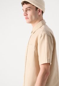 Camicia beige a maniche corte con colletto e tasca frontale, caratterizzata da un logo sottile. Tessuto morbido e leggero con una vestibilità comoda.