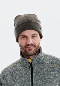 Mörkgrön ribbad stickad mössa med vikbar kant, kombinerad med en texturerad grå zip-up-jacka med en gul accentdragkedja.