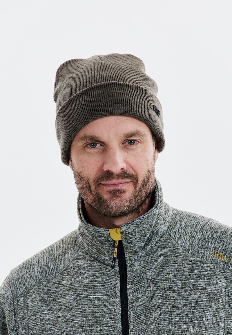 Mörkgrön ribbad stickad mössa med vikbar kant, kombinerad med en texturerad grå zip-up-jacka med en gul accentdragkedja.