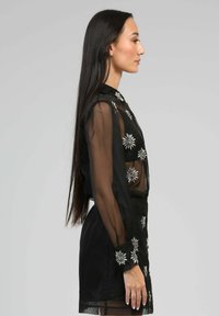 Blouse noire transparente à manches longues, ornée de motifs floraux blancs. Le design comprend une silhouette ajustée et un col montant.