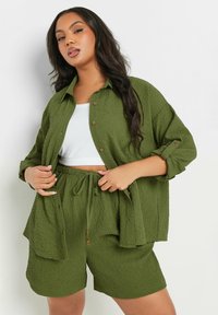 Camicia corta verde oliva con texture e bottoni, abbinata a pantaloni corti con coulisse dello stesso colore. Il tessuto ha un aspetto sgualcito.