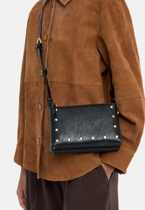 Osoba ubrana w brązową zamszową kurtkę i ciemne spodnie z czarną skórzaną torbą na ramię ozdobioną złotymi ćwiekami.