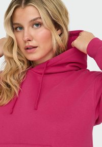 Une femme blonde ajuste la capuche de son hoodie rose vif, regardant légèrement sur le côté contre un fond uni.