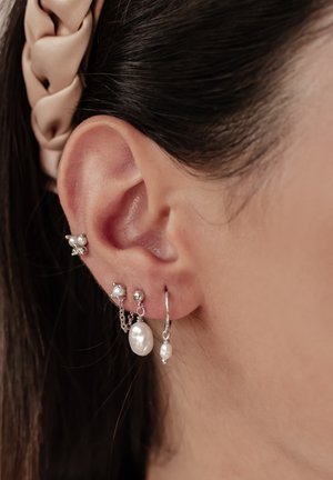 Silberohrringe mit Perlen und einem Kettendesign, die auf einem Ohrläppchen präsentiert werden, im Hintergrund mit einem sanften rosa Stoffhaarbänder.