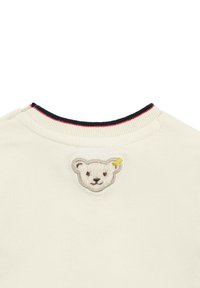Cremfarbendes T-Shirt mit geripptem Ausschnitt, das rote und marineblaue Akzente aufweist. Bestickter Bärenkopf-Patch auf der Rückseite. Weicher Stoff.