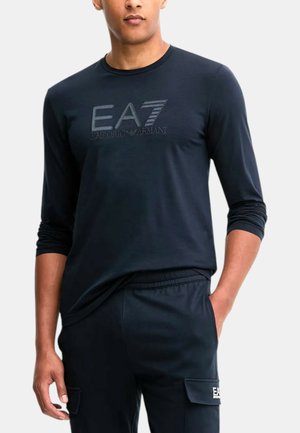 Maglietta a maniche lunghe color blu navy con scollo rotondo, caratterizzata da un logo EA7 testurizzato. Abbinata a pantaloni con vita elasticizzata e tasche cargo dello stesso colore.