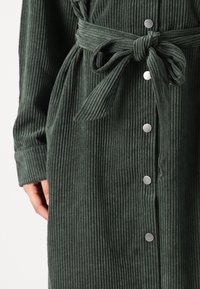 Πράσινο βελούδινο ρούχο με κατακόρυφες ρίγες, που διαθέτει ζώνη, κουμπιά και μανσέτες. Υφα�σμάτινη υφή και μεταλλικά κουμπιά.