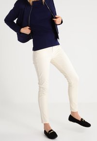 Vita tajta byxor med sidosömdetaljer, kombinerat med en marinblå topp och en svart jacka, kompletterat med svarta loafers med gulddetaljer.