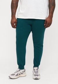 Teal joggerbroeken met elastische boorden en tailleband. Gemaakt van zachte stof. Draag met grijze en witte sneakers.