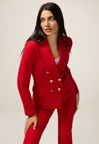 Czerwony garnitur na podwójne guziki z srebrnymi guzikami, dopasowany krój, z bocznymi kieszeniami. Gładka tekstura materiału, stylizowany z białą bluzką pod spodem.
