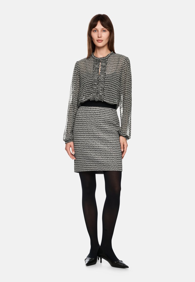 Schwarz-weiße gemusterte Bluse mit gerafftem Ausschnitt, kombiniert mit einem taillierten schwarzen Rock. Model trägt schwarze Strumpfhosen und spitze Schuhe.