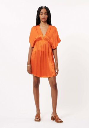Korte oranje satijnen jurk met een diepe V-hals, gathered taille en losse mouwen. Model draagt bruin geklede sandalen en een armband.