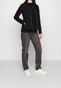 Černá lehká bunda s vysokým límcem a předním zipem, spárovaná s černým svetrem, šedými džíny a bílými teniskami. Jednoduchý design.