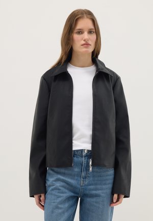 Vrouw met steil haar, gekleed in een zwart jack met rits over een wit shirt en een blauwe spijkerbroek, staand tegen een effen achtergrond.