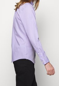 En person i lila och vitrandig långärmad tröja och svarta byxor, sedd från sidan mot en enkel vit bakgrund.