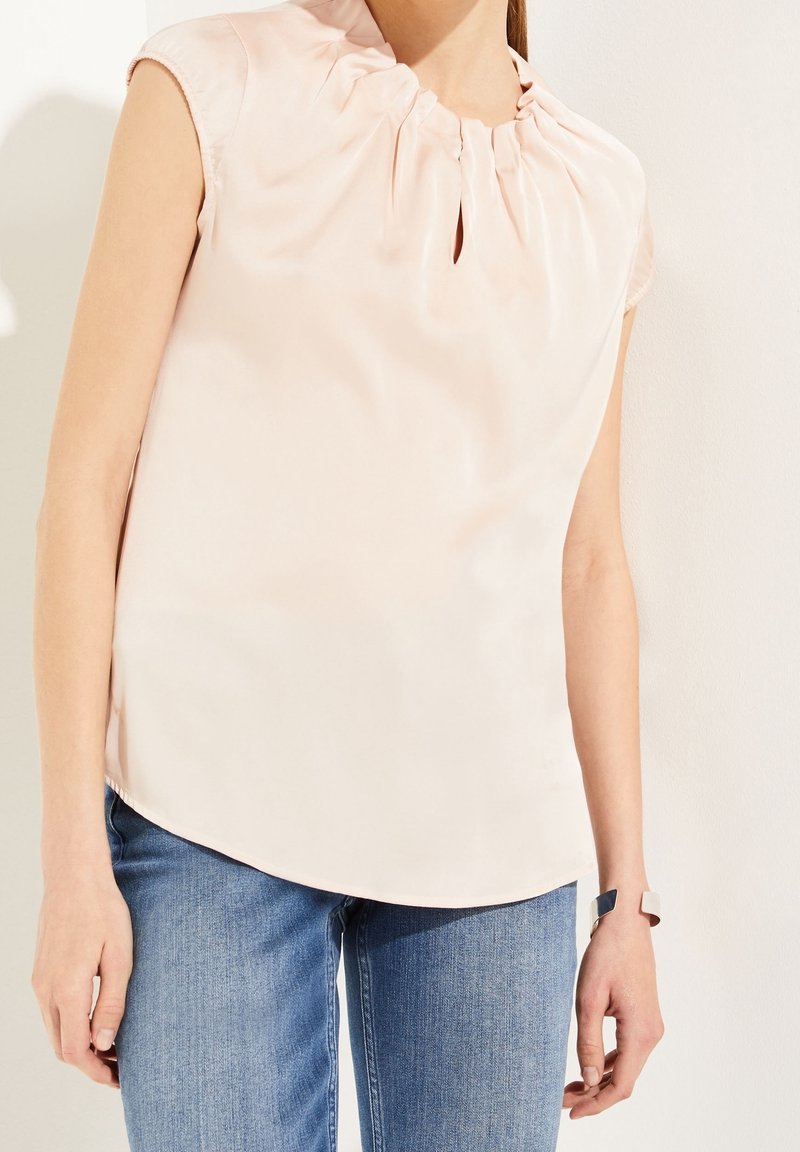 Haut en satin rose clair avec un col plissé et un détail fendu. Manches courtes et texture lisse, porté avec un jean en denim bleu.