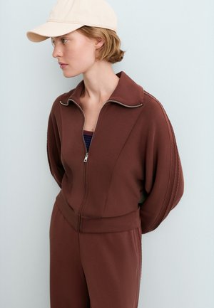 Jeune femme portant une casquette beige et une veste marron zippée avec un pantalon assorti, regardant vers la gauche sur un fond clair uni.