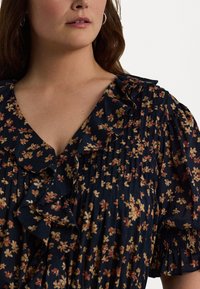 Blomstret kjole med dyp V-hals og volangdetaljer. Har en marineblå bakgrunn med rosa og gule blomster, teksturerte folder og puffede ermer.