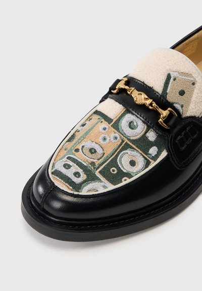 Svart läderloafer med ett överdel i krämfärgad och grön mönstrad textil, prydd med detalj i guldkedja och kontrasterande sömmar.