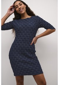 Stickad klänning i mörkblått med ett strukturerat vävt mönster, trekvartsärmar och en figursydd siluett, bärs av en modell mot en neutral bakgrund.