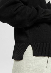 Pull tricoté noir avec ourlet côtelé et fente latérale porté sur un pantalon gris clair, montrant un bras plié et des détails de la texture du tissu.