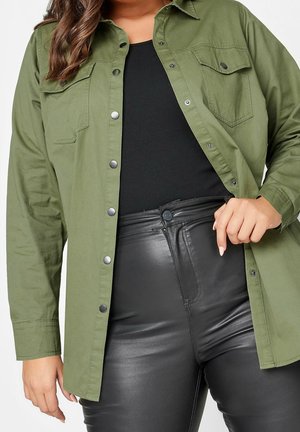 Vrouw draagt een open olijfgroene blouse over een zwarte top, gecombineerd met hooggetailleerde zwarte neplederen broek, hand houdt de zoom van de blouse vast.