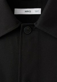 Črna srajca s kragnico in črnimi gumbi, posnetek od blizu, ki prikazuje teksturo tkanine in oznako blagovne znamke MNG z velikostjo EU-USA M, MEX M.