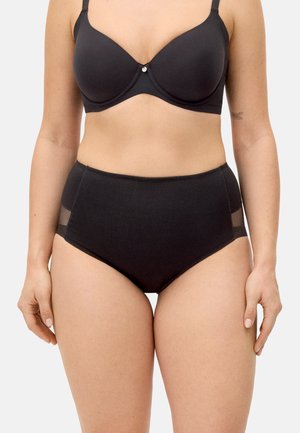 Mujer con sujetador negro con aros y bragas negras de cintura alta con paneles laterales transparantes, de pie contra un fondo liso y claro.