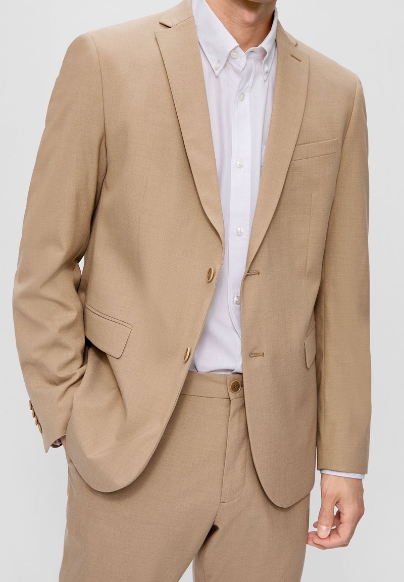 Beige blazer i glat stof, enkeltknappet med to knapper, slagkraver og en brystlomme. Bæres over en hvid skjorte.