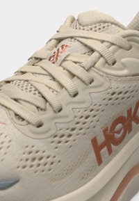 Μπεζ αθλητικό παπούτσι Hoka με διχτυωτό επάνω μέρος, χοντρές κορδόνια και πορτοκαλί λογότυπο "Hoka" στο πλάι και στη γλώσσα.