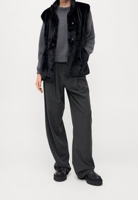 Gilet in pelliccia sintetica nera indossato sopra un maglione grigio lavorato a maglia, abbinato a pantaloni larghi a righe sottili scure e scarpe platform nere e massicce.