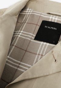 Veste beige avec une texture douce, dotée d'une doublure à carreaux marron et blanc. L'étiquette indique "SCALPERS" en noir.