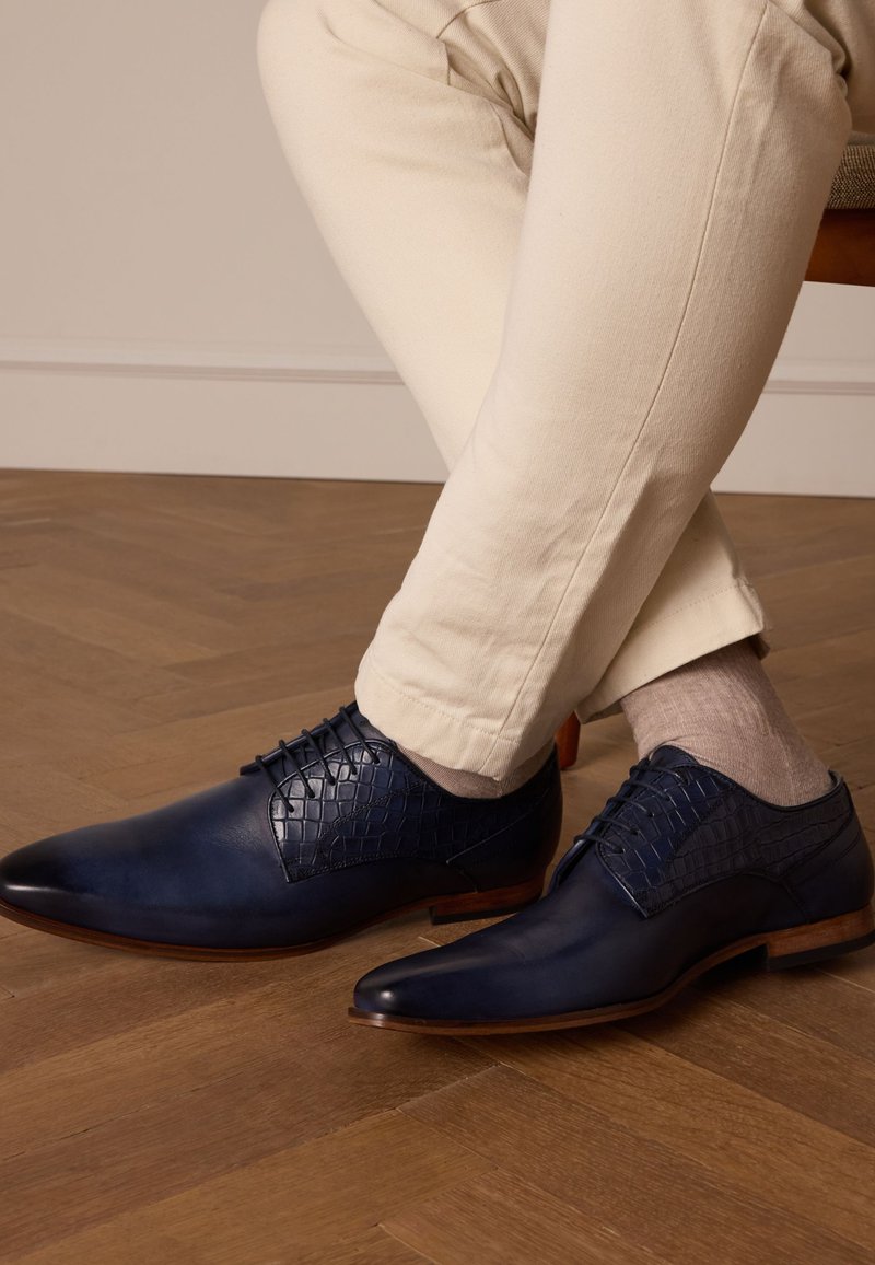 Chaussures en cuir bleu marine avec une texture de crocodile sur le dessus, bout pointu et talons en bois, portées avec un pantalon clair et des chaussettes.