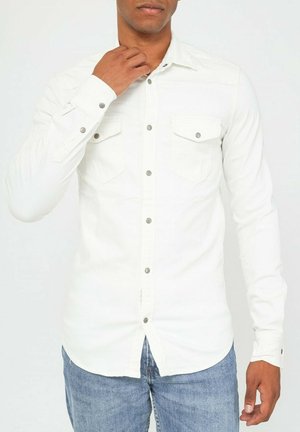 Chemise blanche à manches longues, en coton, avec deux poches poitrine et des accents de boutons en argent, associée à un jean bleu.