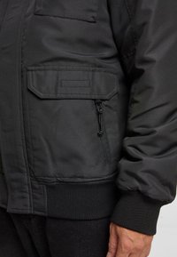 Jachetă bomber din nylon negru, cu un buzunar frontal cu fermoar, manșete din material rib și tiv ribat. Are o textură netedă și un design minimalist.