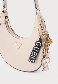 Beżowa skórzana torba na ramię ze złotym metalowym okuciem, trójkątnym logo oraz ozdobnymi zawieszkami przymocowanymi do boku.